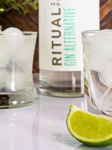 Two glasses of clear liquid with ice cubes and lime slices on a wooden table. a bottle with a label reading "ritual gin alternative" and lime wedges are also visible.