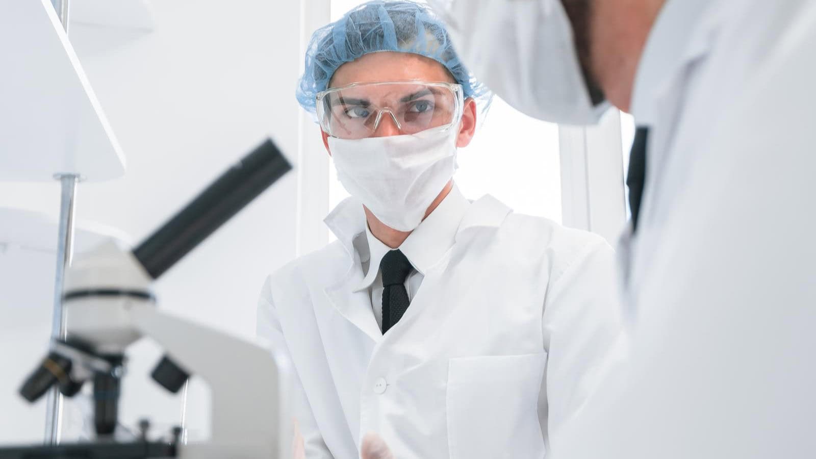 Two scientists in white lab coats and protective gear, including hairnets, masks, and safety goggles, work in a laboratory. One scientist is partially hidden, while the other is in focus near a microscope, suggesting an ongoing experiment or research.