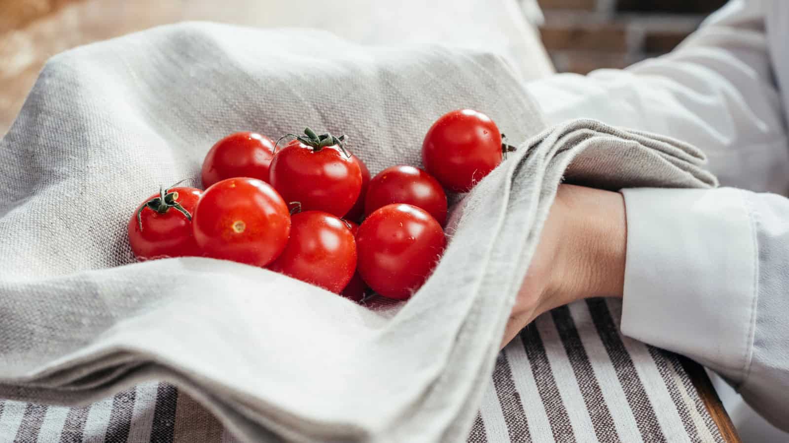 A person holding a bundle of fresh red tomatoes in a grey cloth napkin. The napkin is draped over their hand, with a striped tablecloth visible underneath. The background includes part of a wooden surface and a blurry setting.