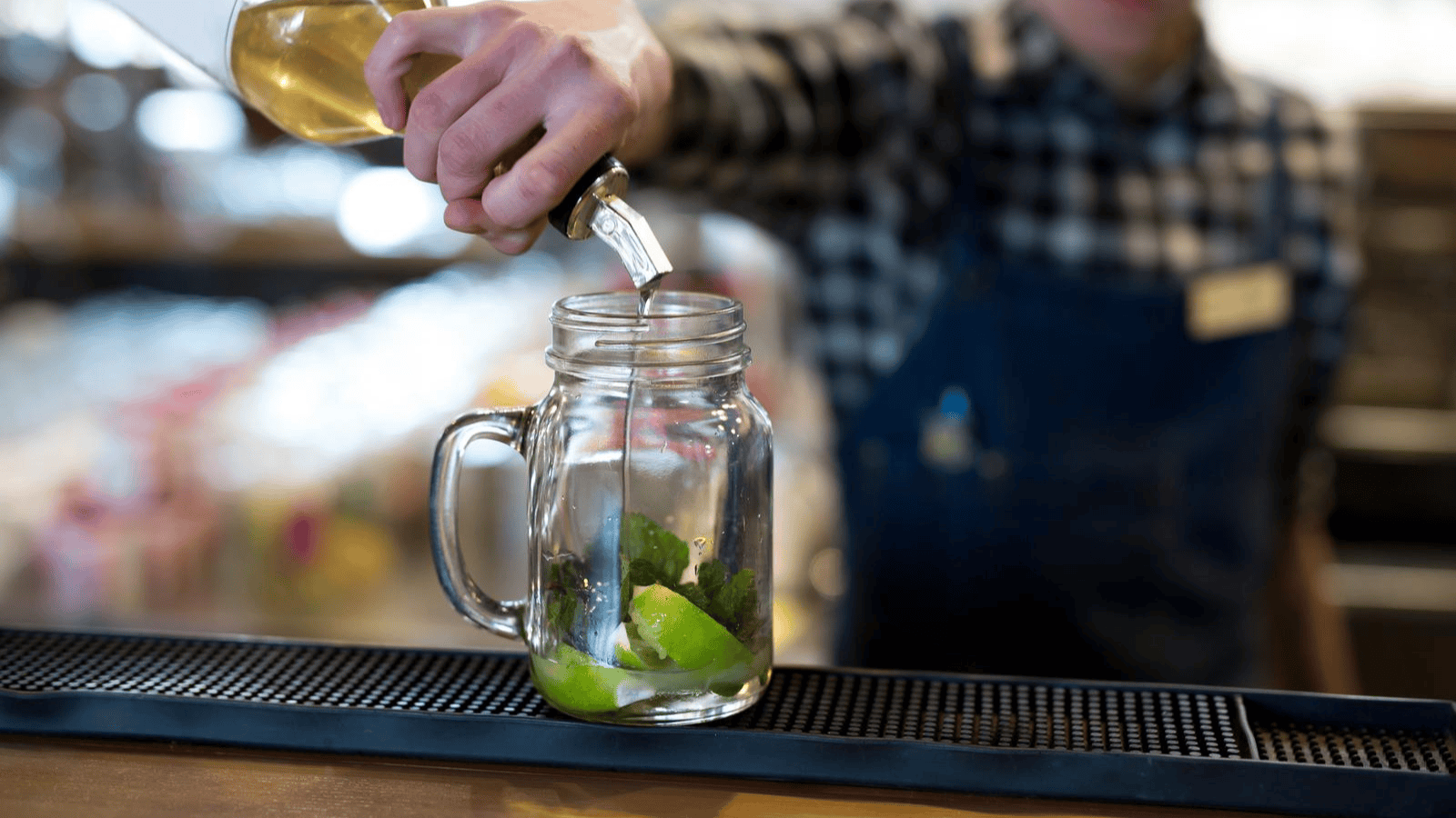 A person wearing a checkered shirt and apron is pouring a liquid from a clear glass bottle into a mason jar containing lime pieces and mint leaves. The jar is on a bar mat, and the background is blurred, showing a bar setting.