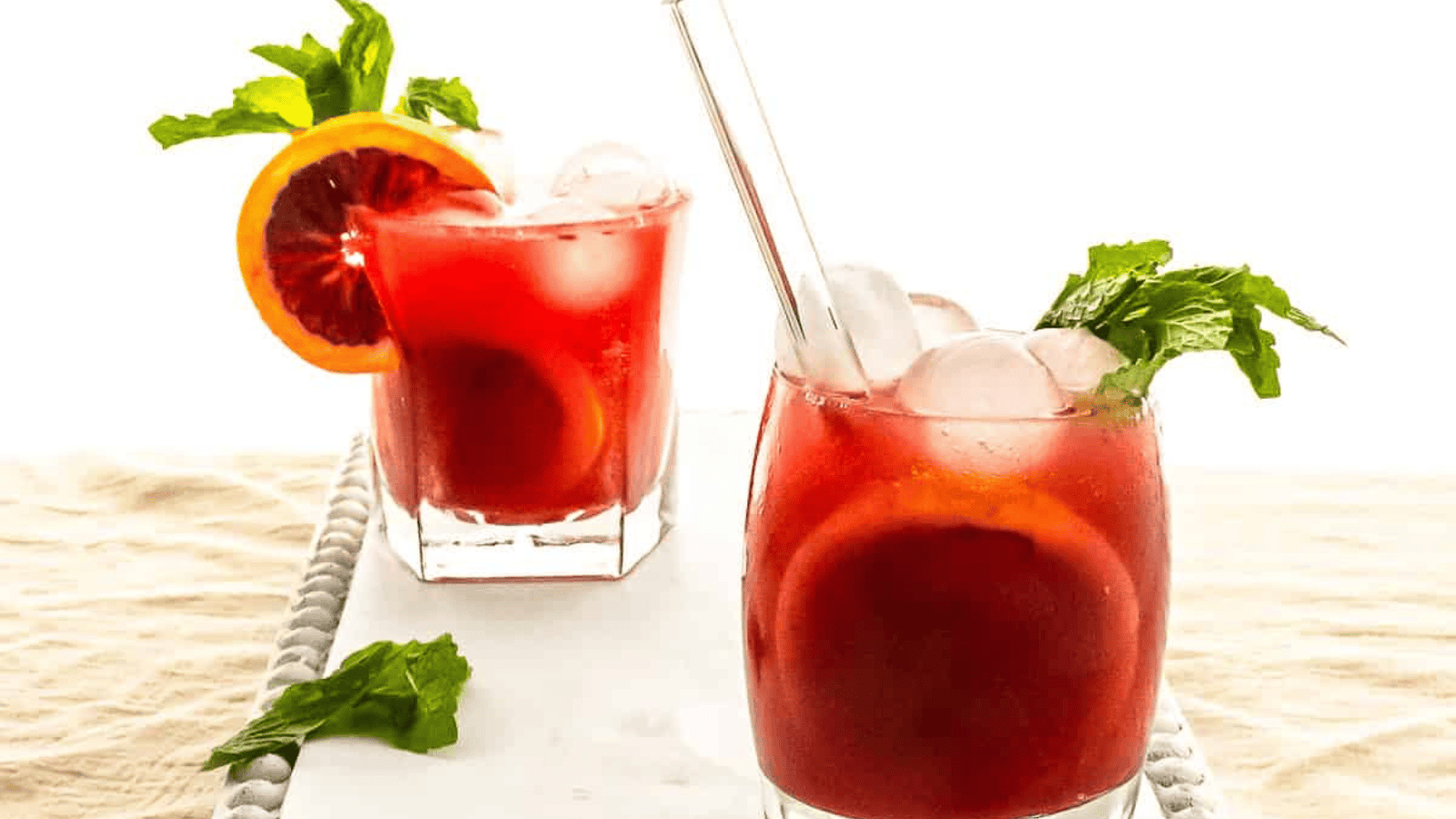 Two glasses of a red beverage with ice are garnished with orange slices and mint leaves. A clear straw is placed in one of the glasses. The drinks are set on a white rectangular tray with a light-colored cloth underneath. Some mint leaves are scattered on the tray.