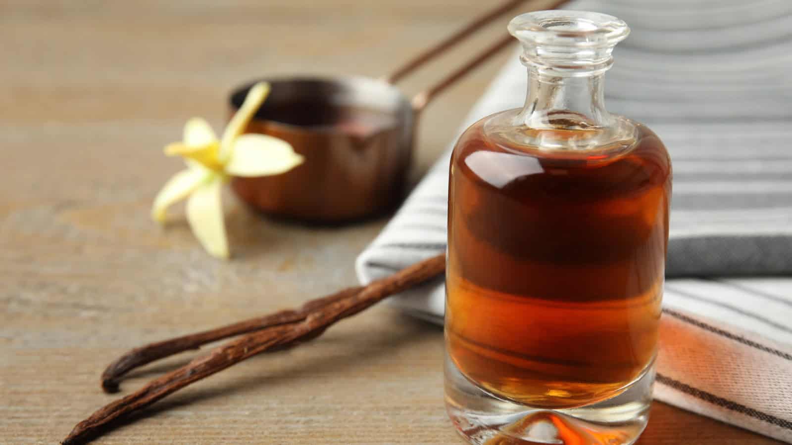 A small glass bottle of vanilla extract is placed on a wooden surface. Next to the bottle are two vanilla beans and a yellow vanilla flower. In the background, there is a metal measuring cup and a folded striped cloth.