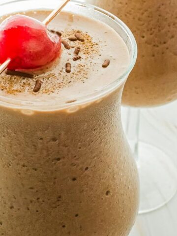 Two glasses of frothy chocolate milkshake topped with a red cherry on a white tabletop.
