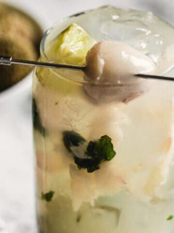 A refreshing cocktail garnished with a lychee and mint leaves, served in a tall glass with ice and a slice of lime, placed on a white surface near a bowl of lychees and fresh mint.