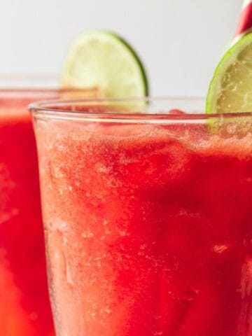 Three glasses filled with bright red watermelon slushies garnished with lime slices, with red and white striped straws, on a white background.