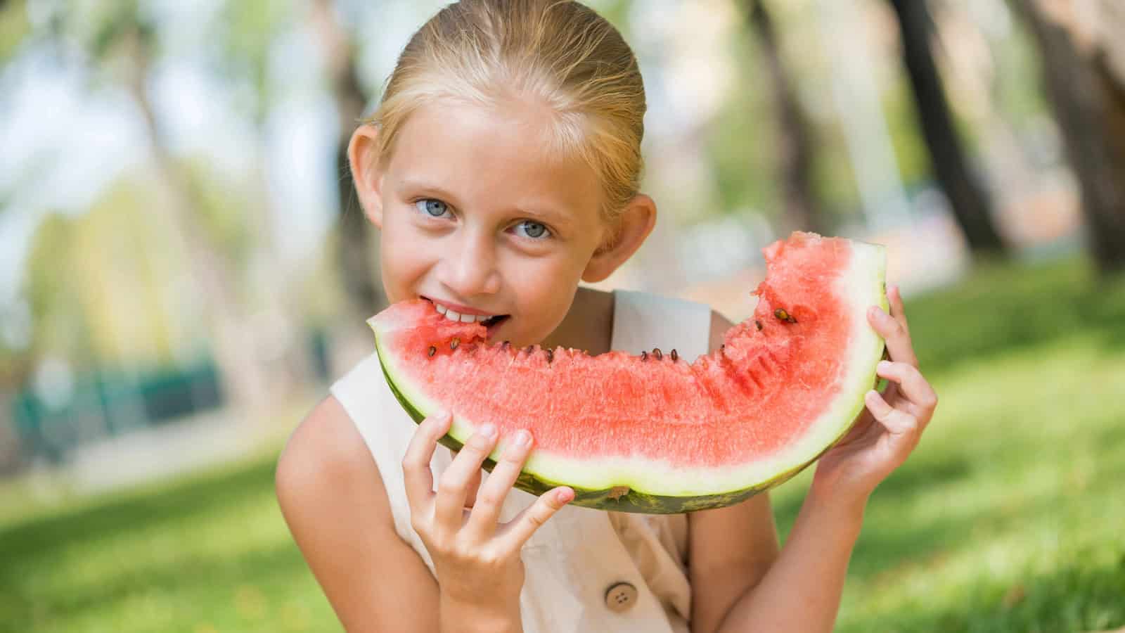 A young girl with blonde hair is sitting on the grass outdoors, holding and eating a large slice of watermelon. She is looking at the camera, and there are blurred trees in the background.