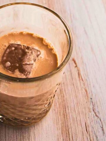 A glass of creamy iced coffee with a square of chocolate floating on top, surrounded by scattered coffee beans on a wooden surface.