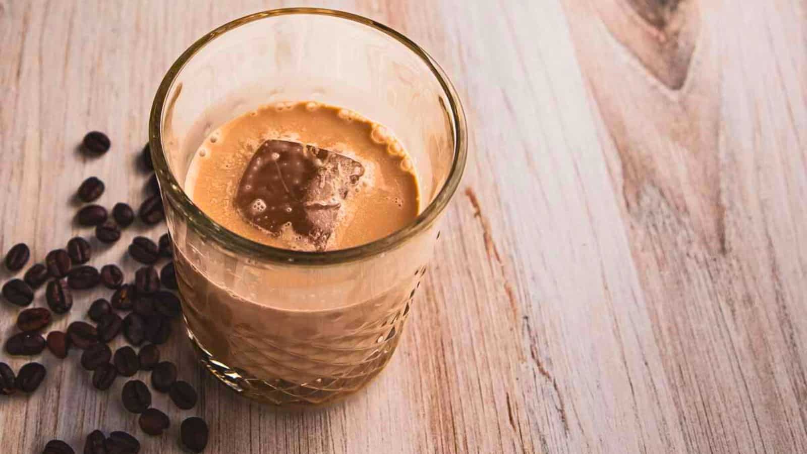 A glass of creamy iced coffee with a square of chocolate floating on top, surrounded by scattered coffee beans on a wooden surface.