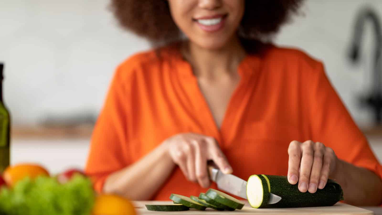 A person wearing an orange shirt is slicing a zucchini with a knife on a wooden cutting board. Other vegetables and an olive oil bottle are blurred in the background.