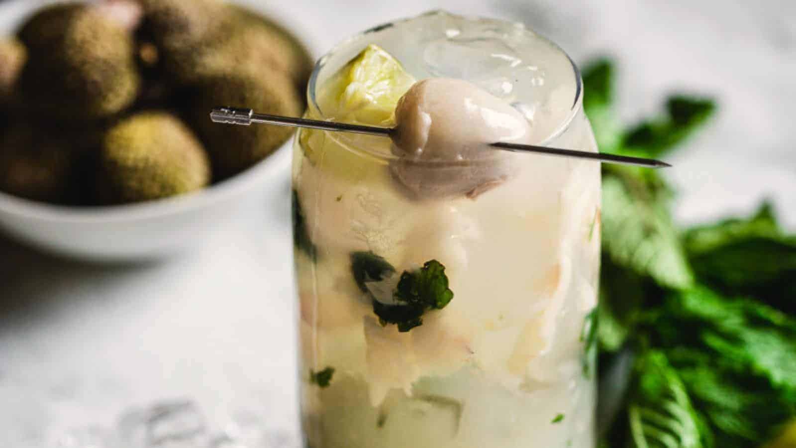 A tall glass filled with a creamy lychee drink garnished with a skewer holding a lychee fruit and a lime slice. Mint leaves and a bowl of lychees are in the background.