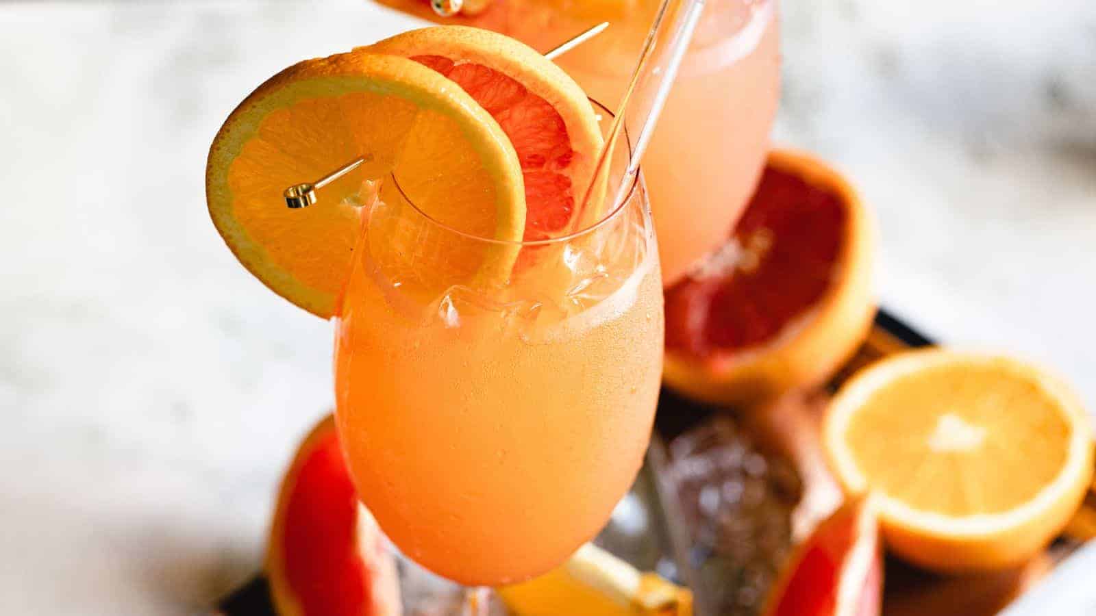 A tall glass filled with a pink-orange beverage, garnished with a slice of orange and a slice of grapefruit on the rim. The background features additional half-cut oranges and grapefruits, and a blurry surface.