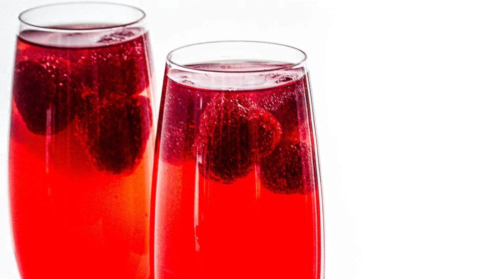 Two glasses filled with a bright red fizzy drink, garnished with floating raspberries, evoke the essence of mocktails that taste like vacation. The background is a plain white, enhancing the vibrant allure of this refreshing escape in a glass.