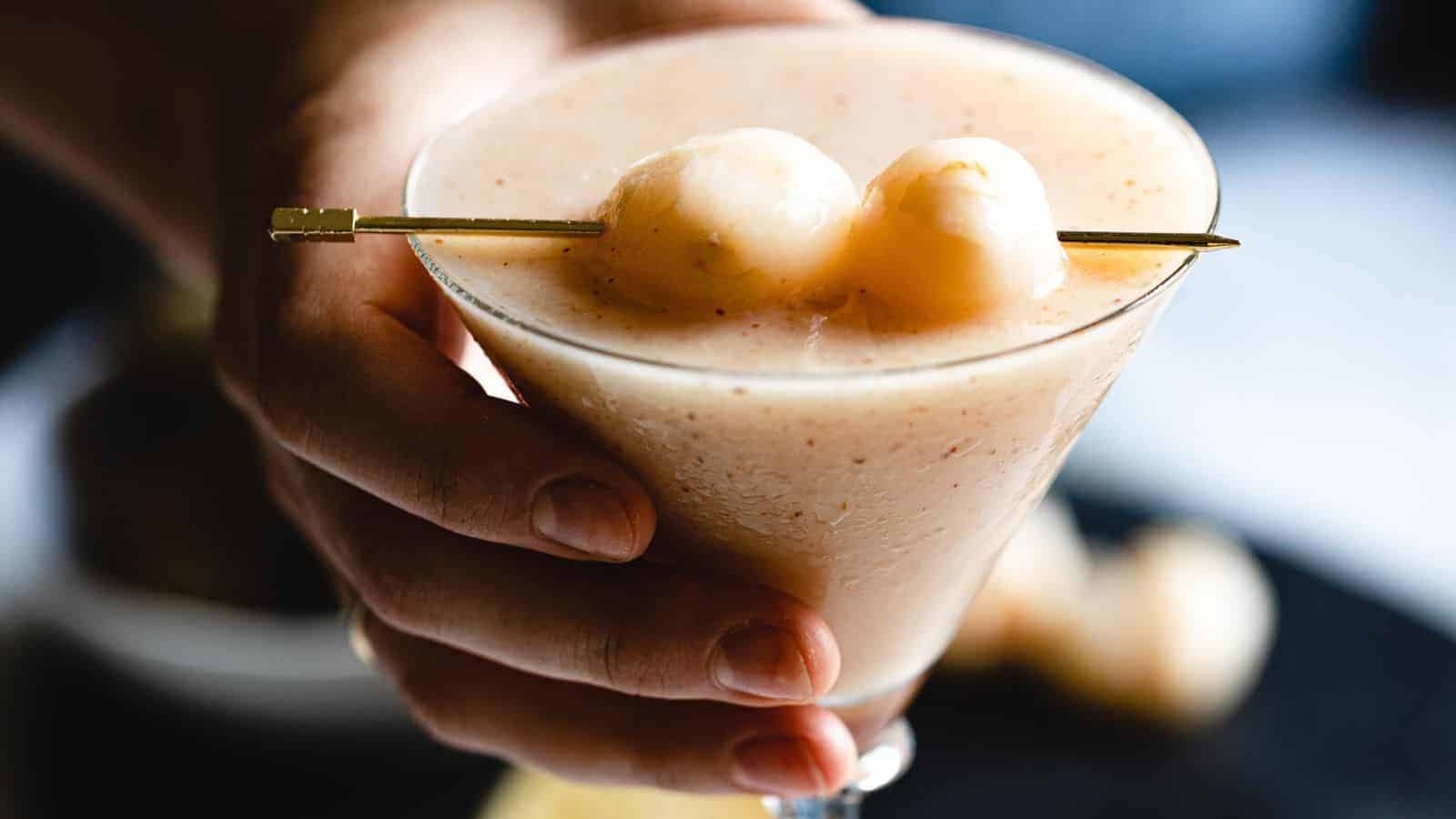 A hand holding a martini glass filled with a light beige frozen cocktail, garnished with two lychee fruits on a skewer. The background is blurred.