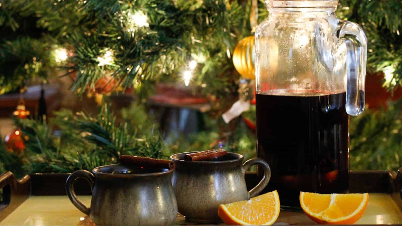 A glass pitcher filled with spiced wine and two mugs of the same beverage sit on a table, accompanied by four orange slices. The background features a decorated and lit Christmas tree, giving a festive atmosphere.