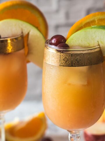 Two glasses of chilled fruit punch garnished with orange slices, apple wedges, and cherries, with a backdrop featuring assorted fruits and a textured white surface.