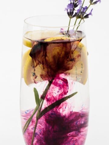 A tall glass filled with clear liquid, slices of lemon, and a small purple flower on top. Dark purple ink is dispersed in the liquid, creating swirling patterns. This artistic drink highlights the beauty of simple syrup substitutes against a plain white background.