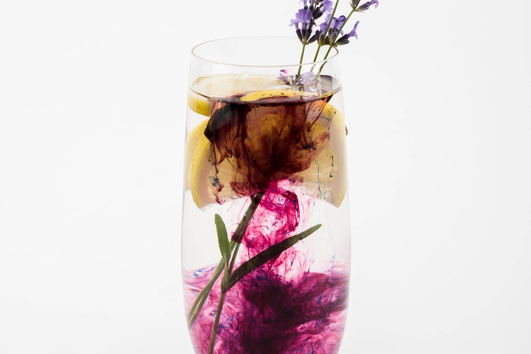 A tall glass filled with clear liquid, slices of lemon, and a small purple flower on top. Dark purple ink is dispersed in the liquid, creating swirling patterns. This artistic drink highlights the beauty of simple syrup substitutes against a plain white background.