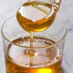 A spoon is shown pouring golden honey, a popular simple syrup substitute, into a clear glass container. The honey appears thick and viscous as it drips from the spoon, creating a smooth, glossy surface in the container below. The background is a neutral, light-colored surface.