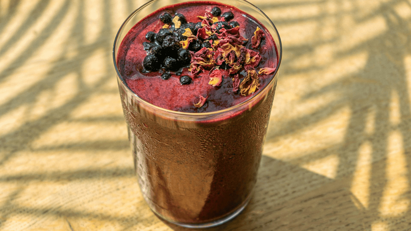A tall glass of superfood smoothie is filled with a dark red blend, topped with blackberries and dried flower petals. It sits gracefully on a wooden surface, where light and shadow dance in harmony.