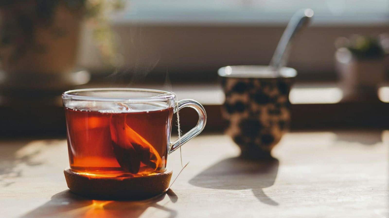A clear glass cup filled with black tea is placed on a wooden surface. A tea bag string is visible hanging from the cup. In the background, there is another out-of-focus cup and some blurred plants, all bathed in natural light.