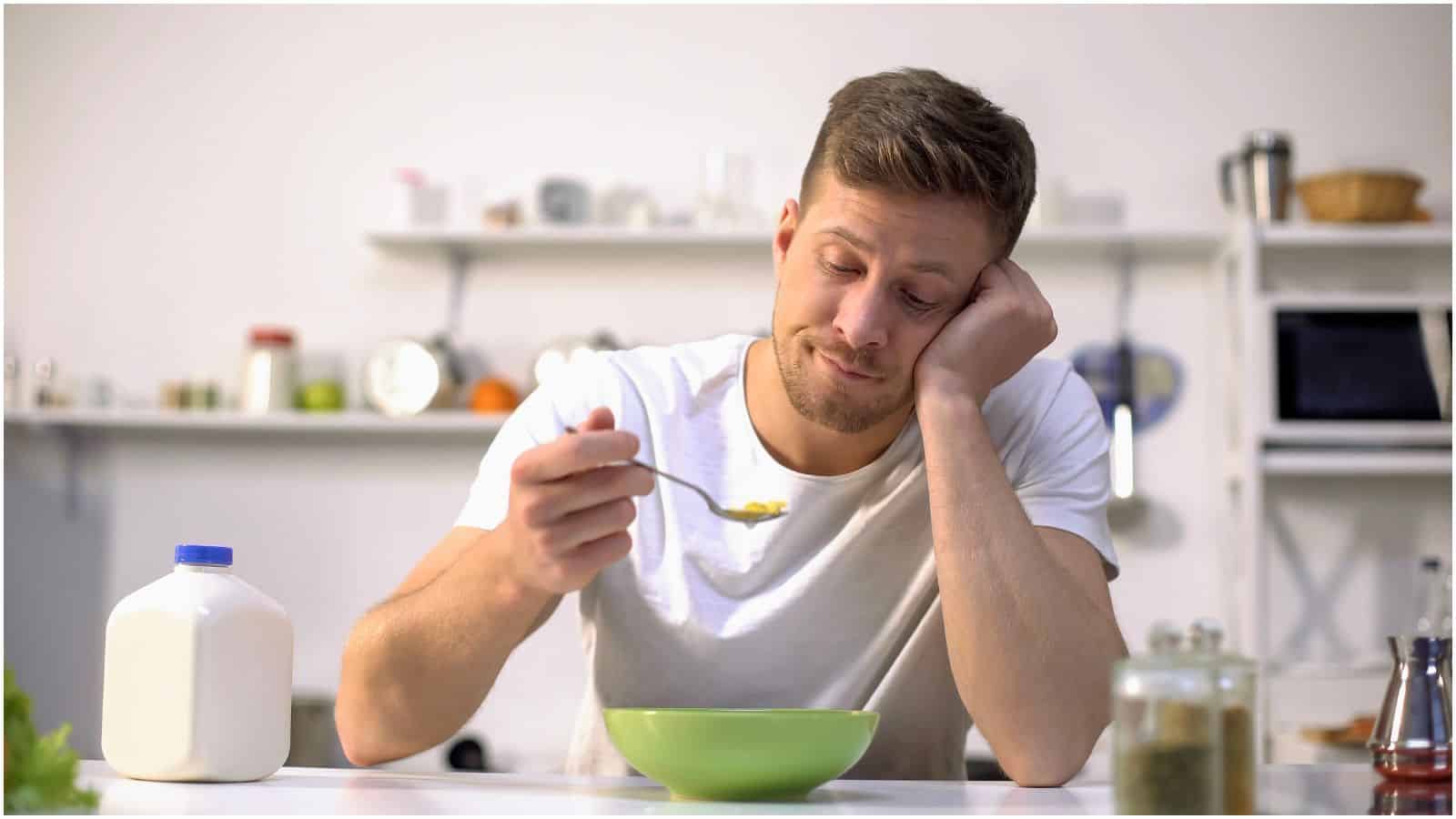 A man in a white t-shirt sits at a kitchen counter with his head resting on one hand. He looks disinterested while holding a spoonful of cereal over a green bowl. A carton of milk is on the left side of the counter, and kitchen shelves are visible in the background.