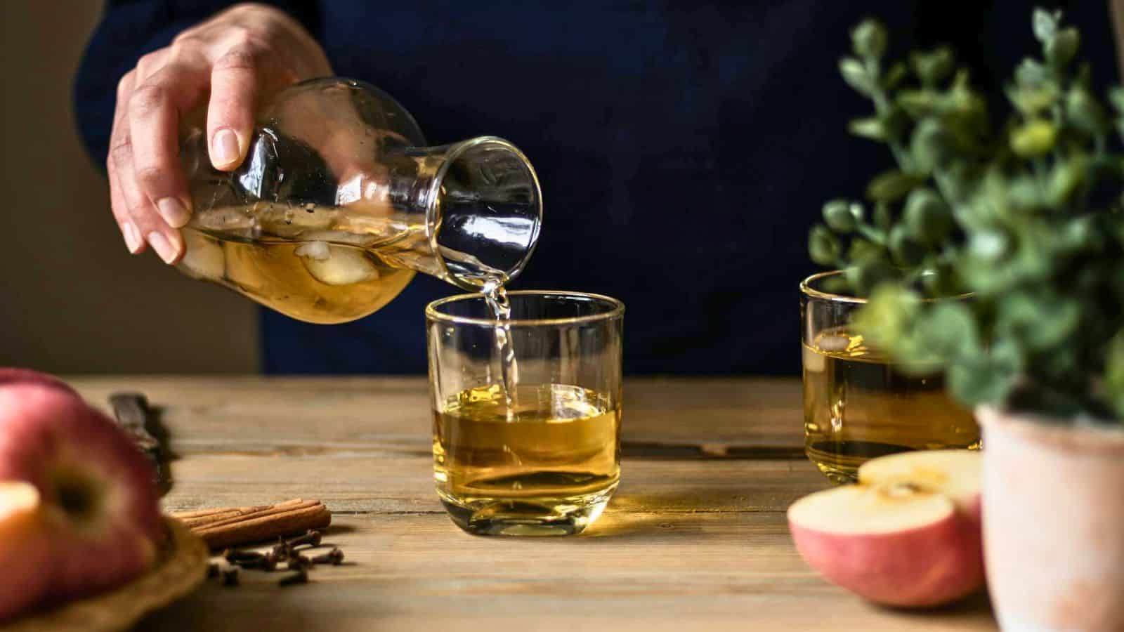 A person pours a light amber liquid from a glass carafe into one of two glasses on a wooden table. The table also features apples, a stick of cinnamon, and a potted plant. The background is dark, contrasting with the lighter items in the foreground.