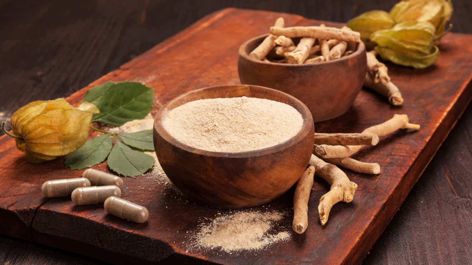 A wooden board displays a bowl of powdered ashwagandha beside a bowl with whole ashwagandha roots. Nearby, there are ashwagandha capsules, leaves, and three greenish-yellow fruit.