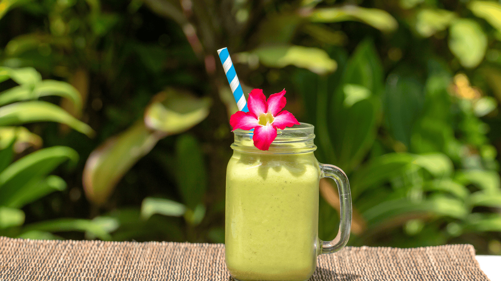 A superfood smoothie in a glass mason jar with a handle, garnished with a red flower and a blue-striped straw, sits on a woven mat. Lush green foliage is blurred in the background.