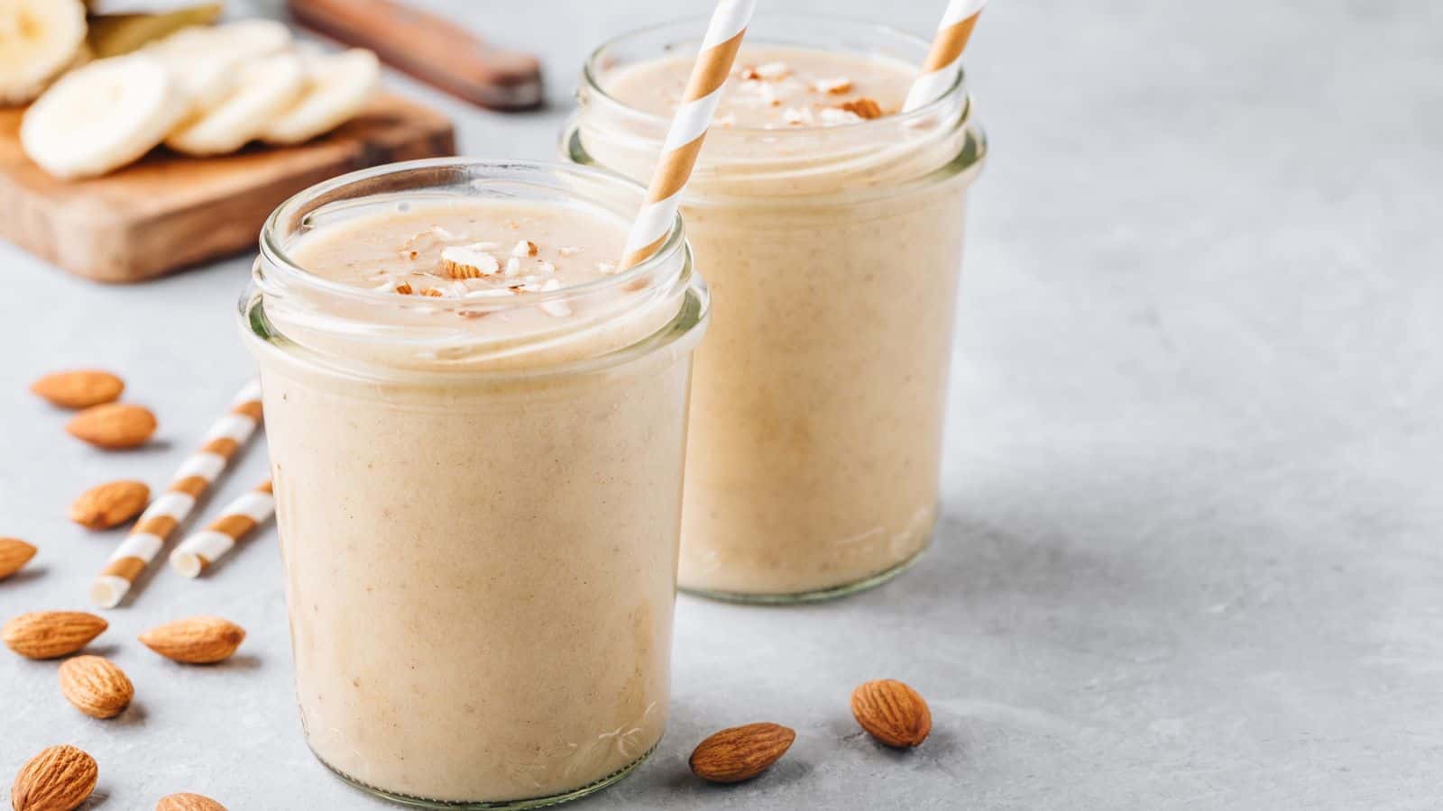 Two glass jars filled with a creamy almond banana smoothie are placed on a light gray surface. Both smoothies are topped with chopped almonds. Each jar has a white and yellow striped straw, while whole almonds and sliced bananas are scattered around.