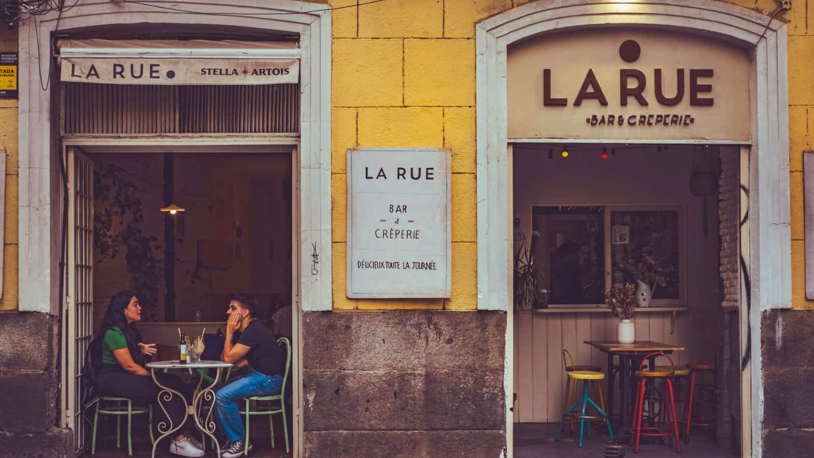 Two people sit at an outdoor table in front of La Rue, a bar and cr&ecirc;perie with yellow walls and stone accents. The sign above the entrance reads "La Rue." Inside, three colorful stools invite more visitors to enjoy this cozy spot.