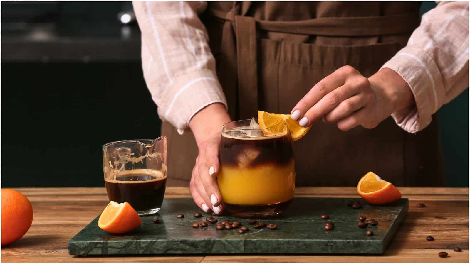 A person in a brown apron is placing an orange slice on the rim of a glass filled with a layered coffee and orange juice drink. Other orange slices, a small jug of espresso, and coffee beans are on the wooden table beside the glass.
