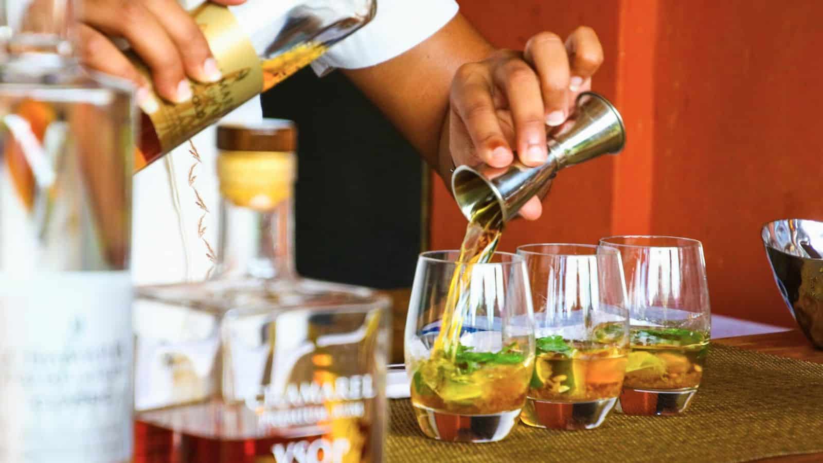 A person is pouring a golden liquid from a jigger into several clear glasses, Bottles of various alcoholic beverages are in the foreground, slightly out of focus. The scene unfolds on a table with a brown mat beneath the glasses.