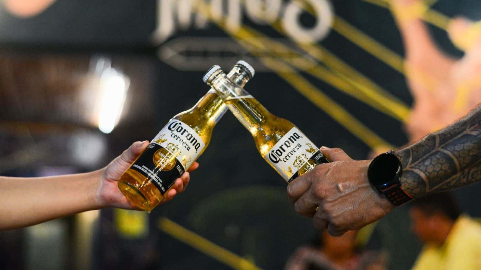 Two people clinking Corona Extra beer bottles together. One person has a visible tattoo and is wearing a wristwatch, while the other person's arm is not fully visible. 