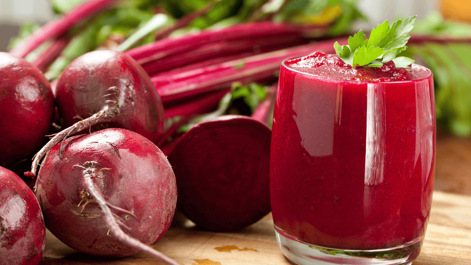 A glass of fresh beet juice, evoking the vibrancy of superfood smoothies, is garnished with a parsley leaf on a wooden surface. In the background, several whole beets with leafy stems complete this nutritious scene.