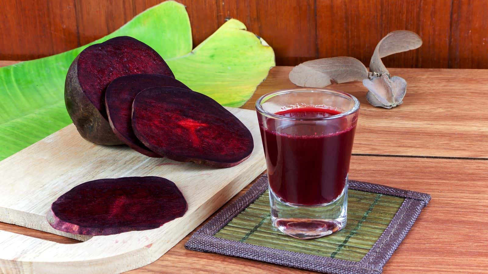 A glass of red juice sits on a coaster on a wooden table. Beside it, a cutting board holds slices of dark red fruit with more slices placed around. A green leaf, brown seed pod, and wooden back wall complete the scene.
