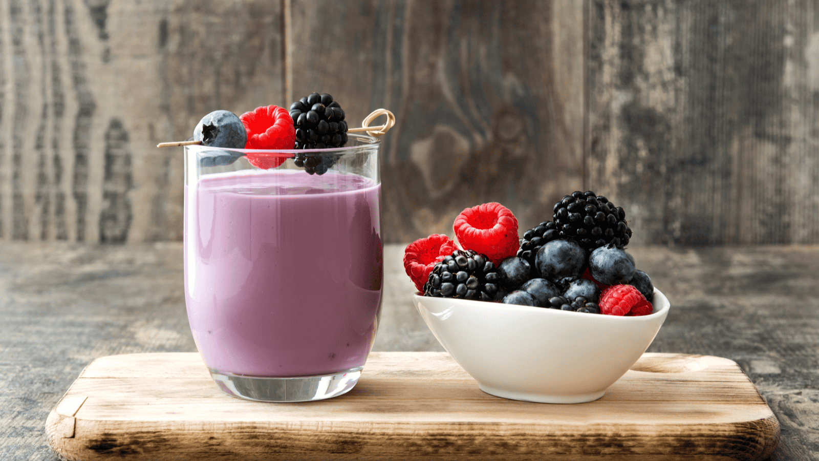 A glass of superfood berry smoothie sits on a wooden board, garnished with a skewer of blueberries, raspberries, and blackberries. Next to it, a white bowl brims with assorted fresh berries. The background is rustic wood.