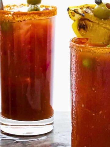 Two glasses of Bloody Mary cocktails are garnished with olives, a pickle spear, and a lemon wedge. A bottle of Lea & Perrins Worcestershire Sauce and a bottle of Tabasco hot sauce are placed next to the glasses on a wooden surface.