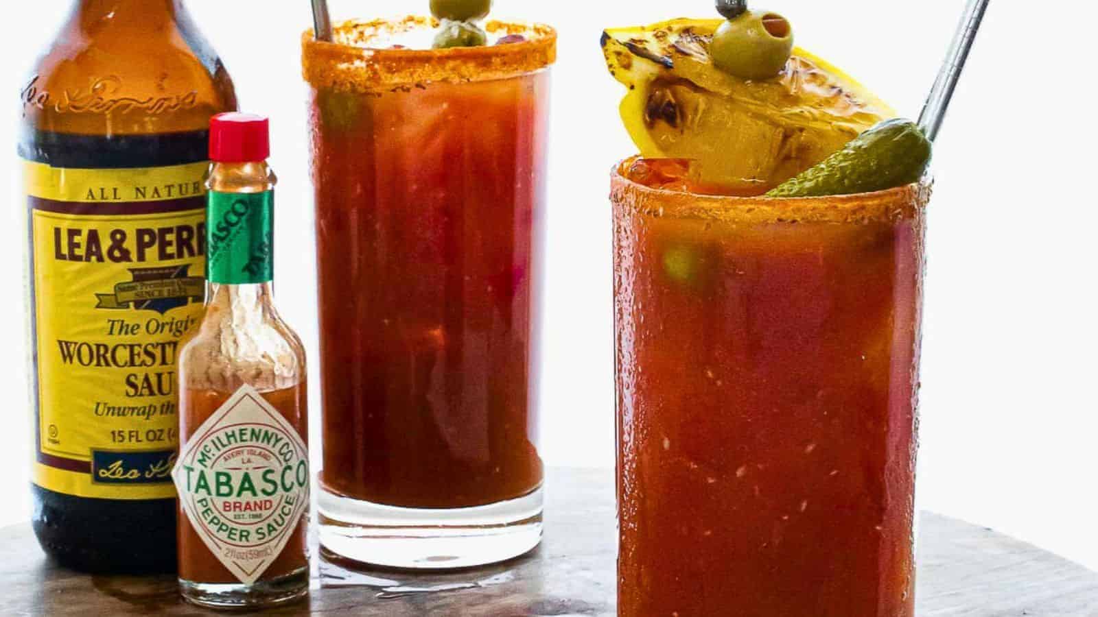 Two glasses of Bloody Mary cocktails are garnished with olives, a pickle spear, and a lemon wedge. A bottle of Lea & Perrins Worcestershire Sauce and a bottle of Tabasco hot sauce are placed next to the glasses on a wooden surface.