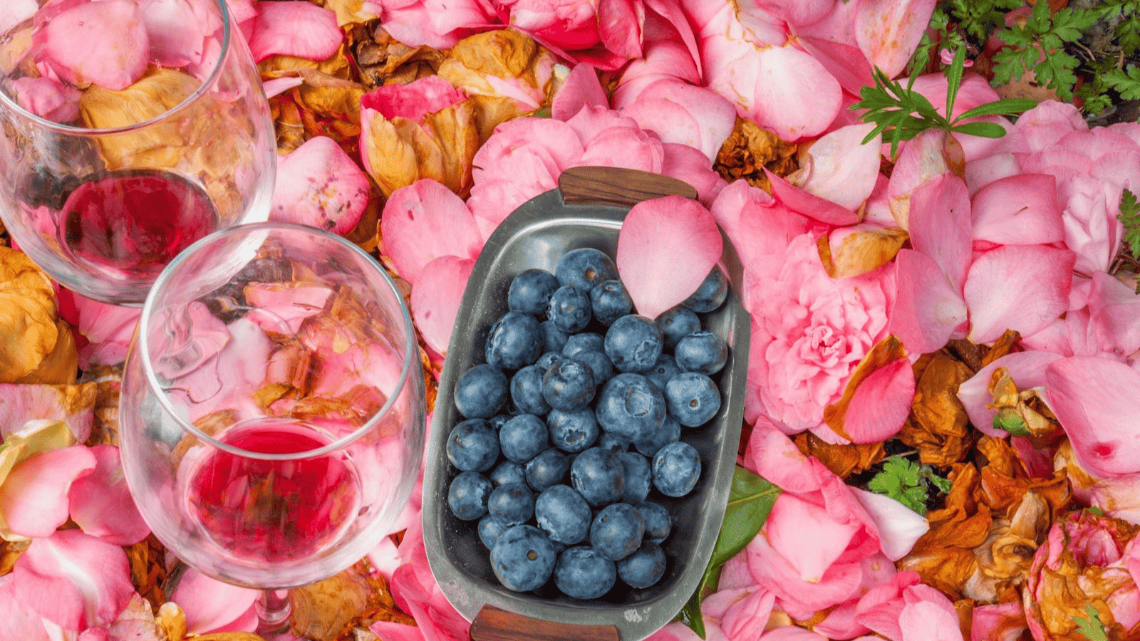A metal tray of blueberries rests on a bed of pink rose petals, with two glasses of red wine nearby. The vibrant scene combines the rich hues of blueberries and wine with the soft pink petals, an homage to unusual plants used for winemaking.