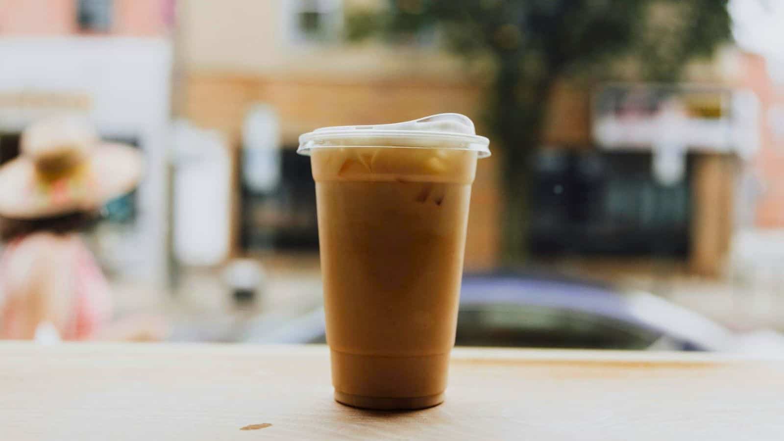 A plastic cup filled with iced coffee is placed on a light-colored wooden table. The background showcases a blurred view of a street with buildings, a tree, and a person wearing a hat.