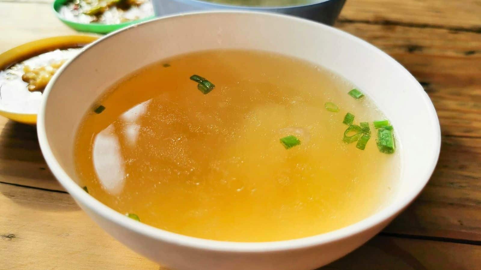 A white bowl filled with clear broth, garnished with chopped green onions, sits on a wooden table. It stands out in the foreground while two other bowls with different contents are slightly blurred in the background.