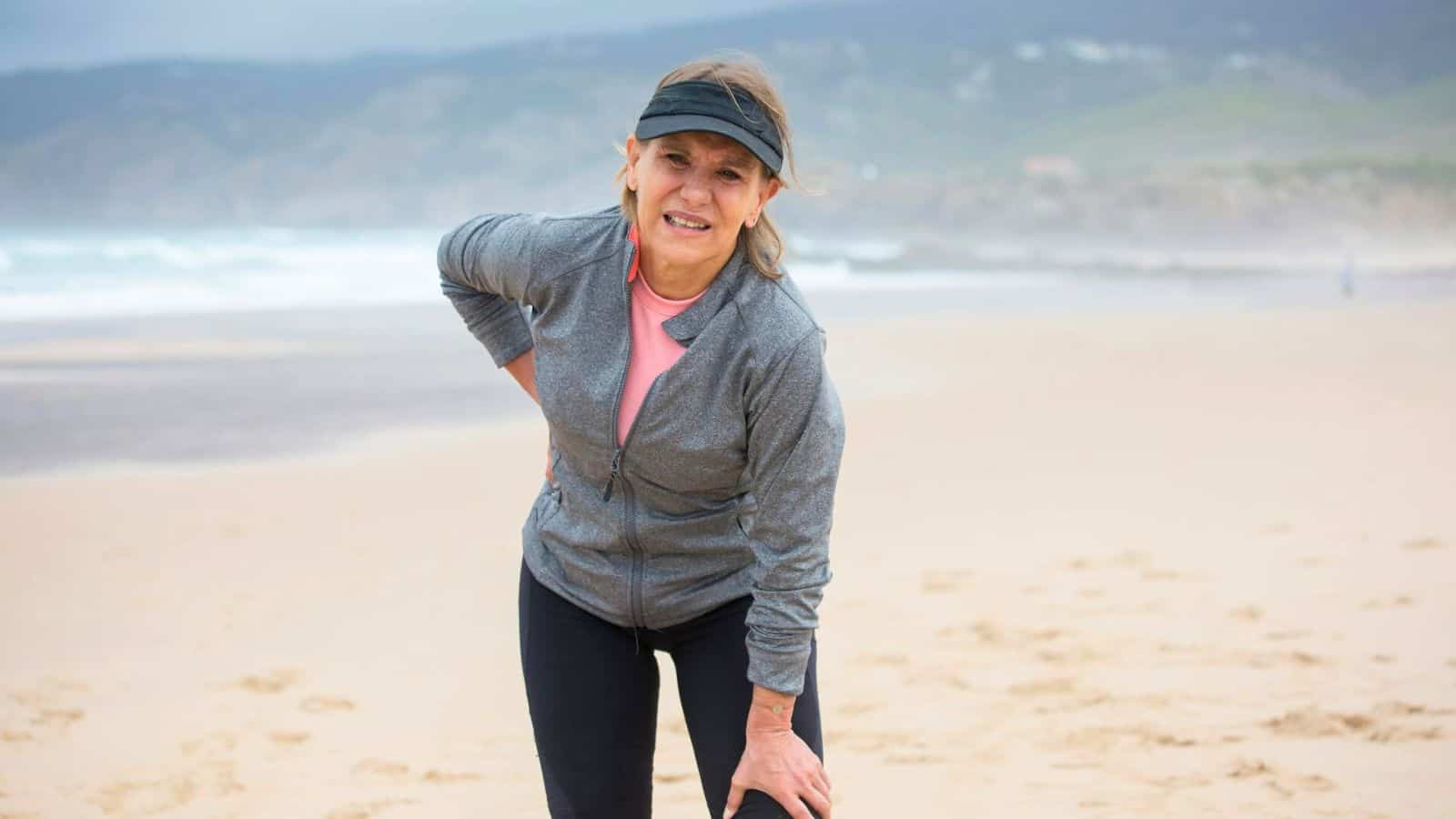 An older woman wearing a visor, a gray zip-up jacket, and black athletic pants is standing on a beach. She is leaning forward slightly with one hand on her knee and the other on her hip. 