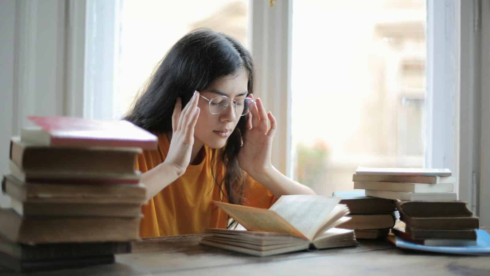 A person with long hair and glasses is sitting at a table by a window, holding their head with both hands and looking down at an open book. There are several stacks of books on the table around them.