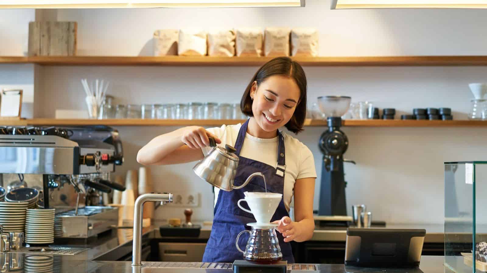 A woman wearing a blue apron and a white t-shirt is pouring hot water from a kettle into a pour-over coffee dripper at a coffee shop. She is smiling and standing behind a counter with a coffee machine, cups, and shelves filled with items.