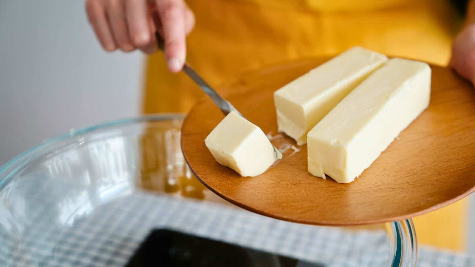 A person in a yellow apron is slicing a stick of butter on a wooden cutting board. The cutting board holds two additional sticks of butter, and a glass mixing bowl sits on a checkered surface underneath.