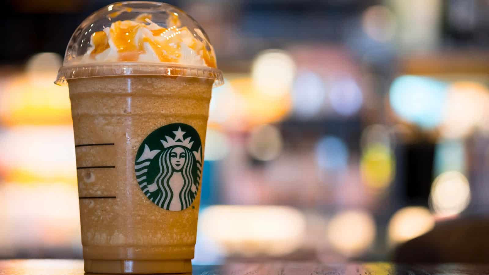 A Starbucks caramel frappuccino with whipped cream and a straw is placed on a wooden table. The background is blurred, showing an indoor setting with warm lighting, hinting at the cozy ambiance.