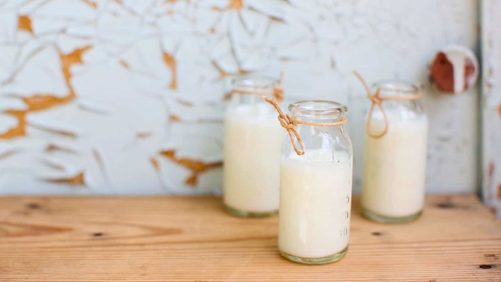 Three small glass bottles filled with a white liquid, likely buttermilk, are placed on a wooden surface. Each bottle is tied with a string around the neck. The background includes a faded white textured wall with peeling paint.