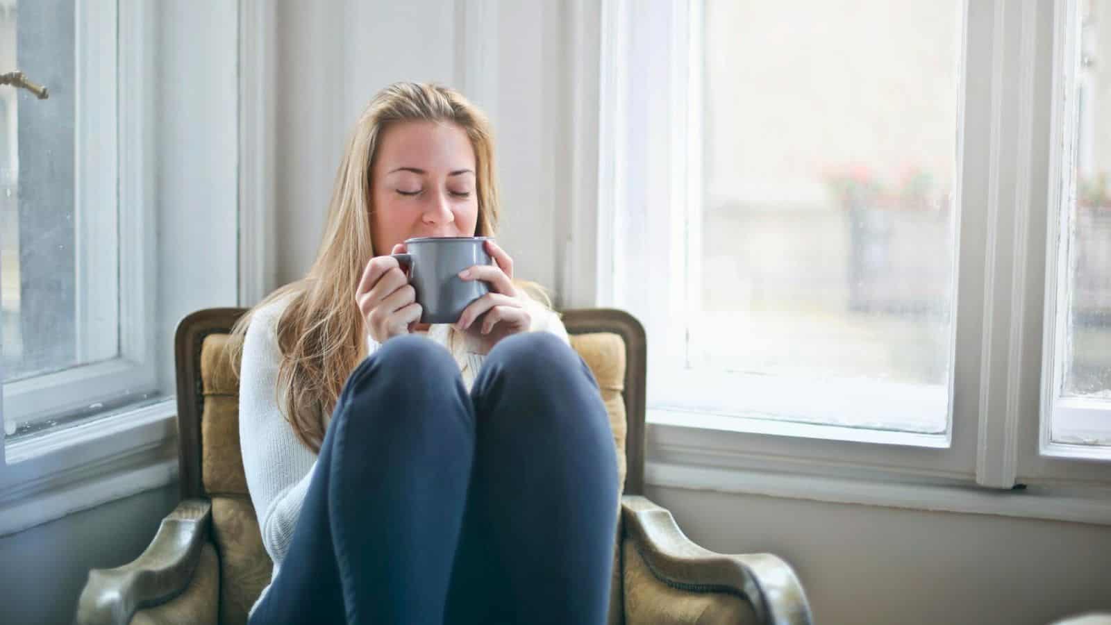A woman with long blonde hair is sitting in a cozy chair by the window, holding a gray mug with both hands. She is wearing a light gray sweater and navy blue pants and appears to be enjoying tea with her eyes closed.