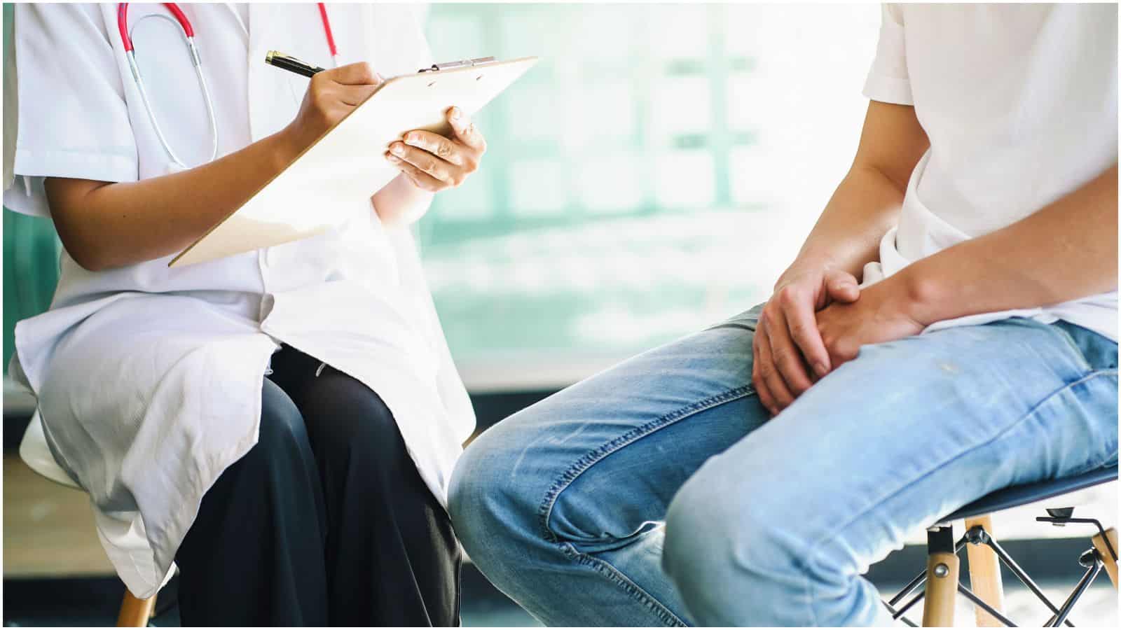 A healthcare professional wearing a white coat and a stethoscope is holding a clipboard, writing on it. Next to them, a person in a white shirt and blue jeans sits on a stool. 