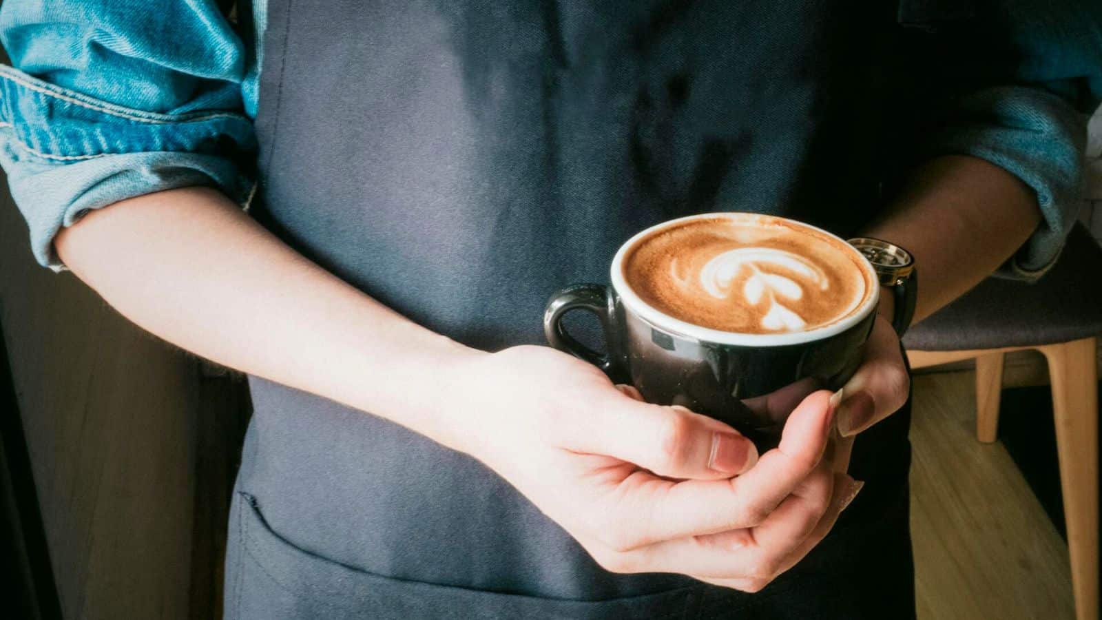 A person wearing a denim shirt and dark apron holds a black cup of cappuccino with latte art. The coffee has a fern-like design on its cream.